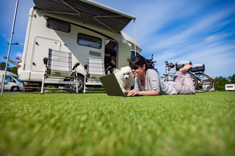 Woman lying on the grass with a dog looking at a laptop. Pet friendly RV rental in background